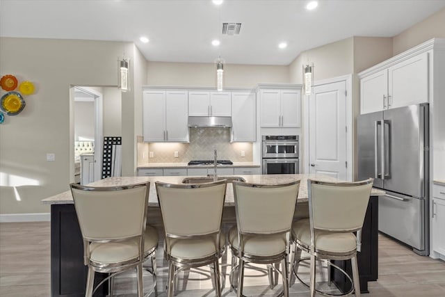 kitchen with white cabinetry, a kitchen island with sink, decorative backsplash, appliances with stainless steel finishes, and light wood-type flooring