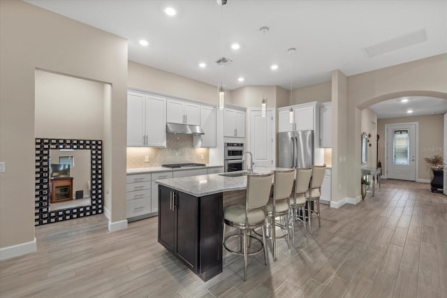 kitchen featuring appliances with stainless steel finishes, light wood-type flooring, pendant lighting, white cabinetry, and an island with sink