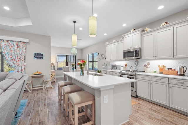 kitchen with light wood-type flooring, stainless steel appliances, sink, decorative light fixtures, and an island with sink