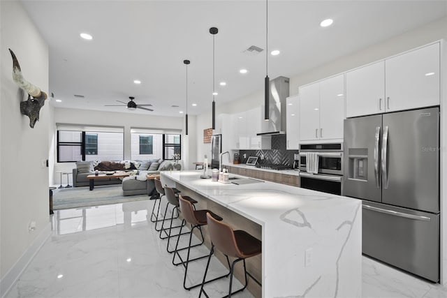 kitchen with stainless steel fridge, wall chimney range hood, pendant lighting, a center island with sink, and white cabinetry