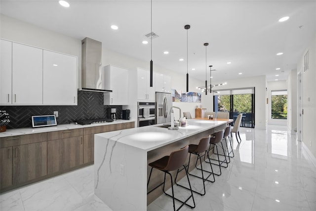 kitchen with white cabinetry, wall chimney exhaust hood, an island with sink, pendant lighting, and appliances with stainless steel finishes