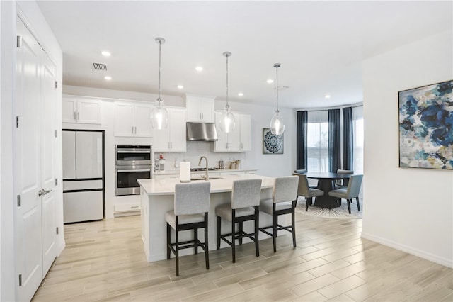 kitchen with white cabinetry, white refrigerator, double oven, an island with sink, and pendant lighting