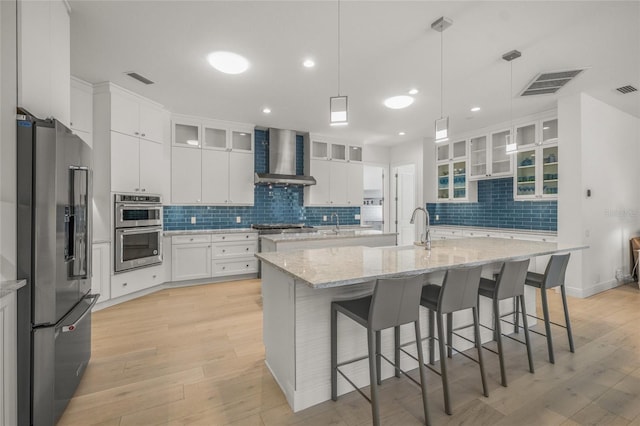 kitchen with stainless steel appliances, a kitchen island with sink, wall chimney range hood, white cabinets, and light hardwood / wood-style floors