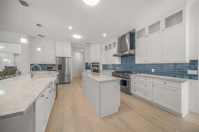 kitchen with wall chimney exhaust hood, stainless steel appliances, a kitchen island with sink, white cabinets, and light hardwood / wood-style floors