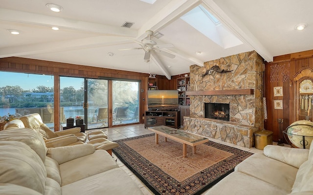 living room featuring a stone fireplace, wood walls, light tile patterned floors, ceiling fan, and vaulted ceiling with skylight