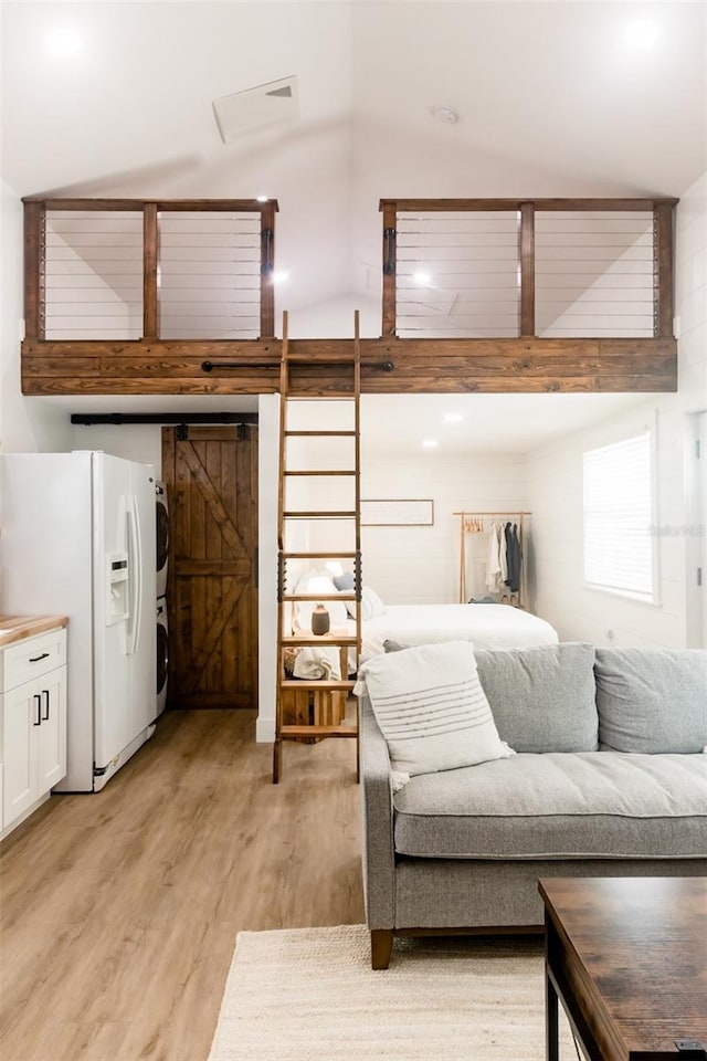 bedroom with white fridge with ice dispenser, a barn door, light hardwood / wood-style flooring, stacked washer and dryer, and vaulted ceiling