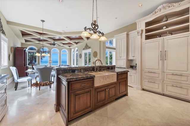 kitchen with coffered ceiling, sink, hanging light fixtures, dark stone countertops, and an island with sink