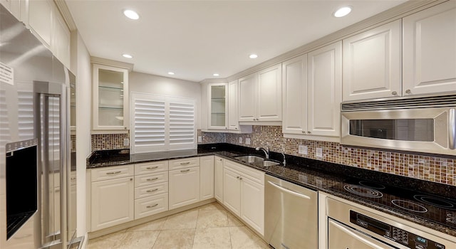 kitchen with stainless steel appliances, a sink, backsplash, dark stone counters, and glass insert cabinets