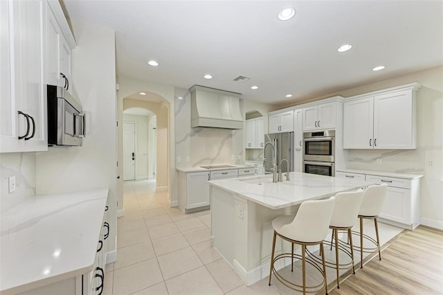 kitchen featuring light stone countertops, appliances with stainless steel finishes, premium range hood, a center island with sink, and white cabinetry