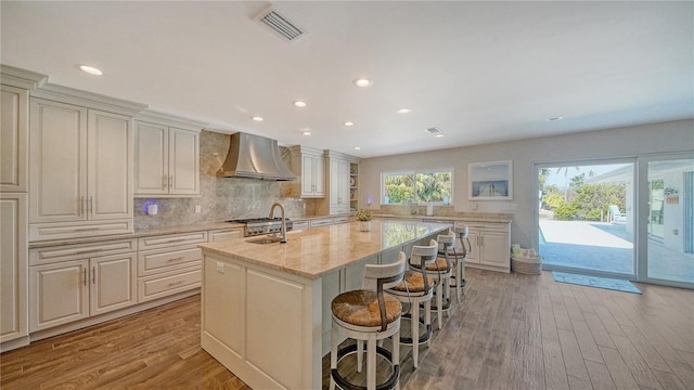 kitchen featuring a center island with sink, wall chimney exhaust hood, light stone countertops, tasteful backsplash, and a breakfast bar area