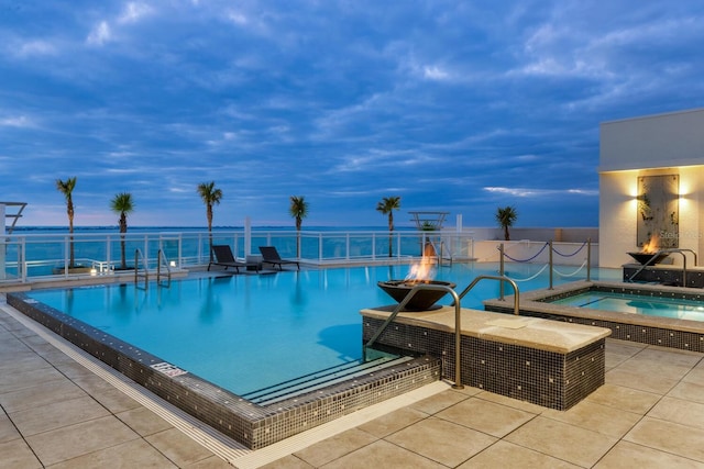 pool at dusk featuring a patio area, a water view, a hot tub, and an outdoor fire pit