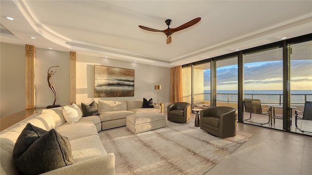 living room featuring a raised ceiling, ceiling fan, a water view, and a wall of windows