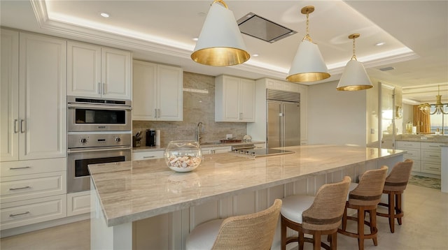 kitchen with light stone counters, stainless steel appliances, a raised ceiling, a spacious island, and hanging light fixtures