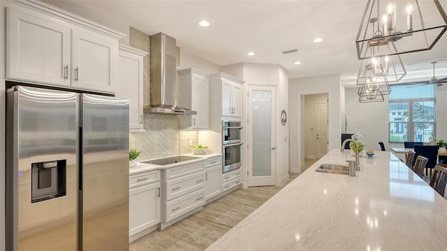 kitchen with sink, wall chimney range hood, decorative light fixtures, white cabinets, and appliances with stainless steel finishes