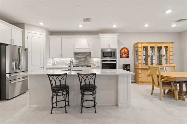 kitchen with custom exhaust hood, white cabinetry, stainless steel appliances, and a kitchen island with sink