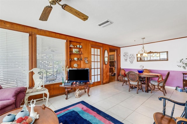 tiled living room with ceiling fan with notable chandelier, built in features, ornamental molding, and wooden walls