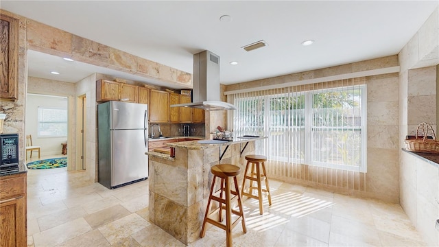 kitchen featuring a center island, a breakfast bar area, light stone countertops, island exhaust hood, and stainless steel refrigerator