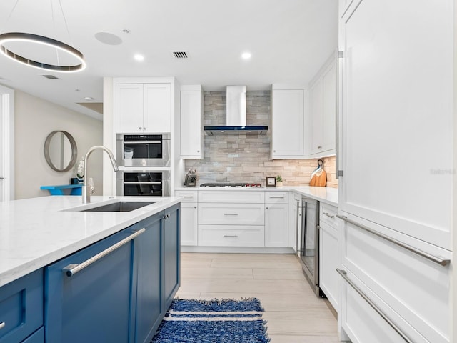 kitchen with pendant lighting, wall chimney range hood, sink, blue cabinetry, and white cabinets