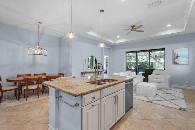 kitchen with a tray ceiling, ceiling fan, sink, decorative light fixtures, and an island with sink