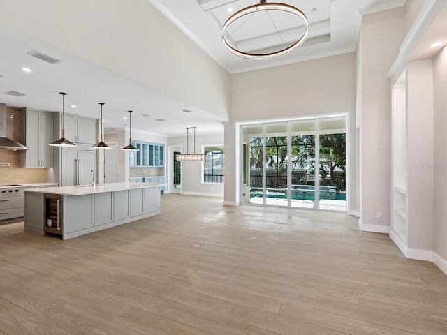 kitchen with gray cabinetry, a kitchen island with sink, beverage cooler, wall chimney range hood, and hanging light fixtures