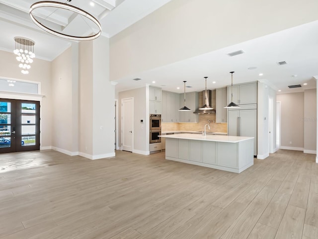 kitchen featuring sink, stainless steel double oven, wall chimney range hood, a notable chandelier, and a kitchen island with sink