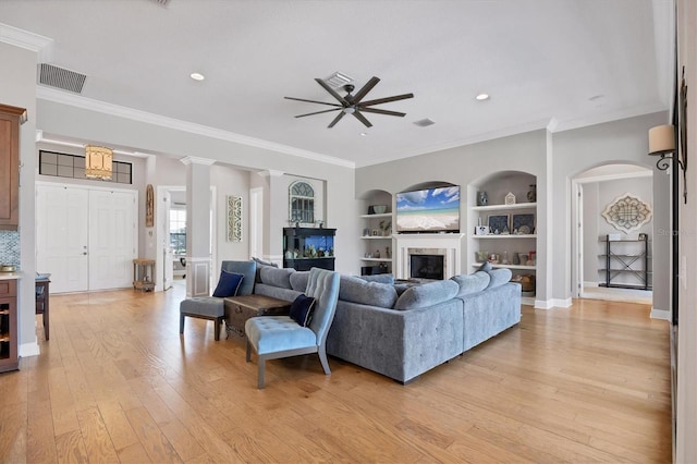 living room featuring built in shelves, ceiling fan, light wood-type flooring, ornamental molding, and decorative columns