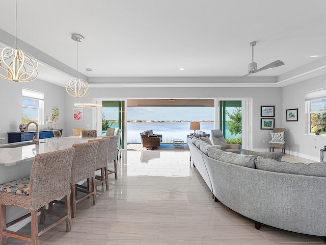 living room featuring ceiling fan with notable chandelier, a water view, sink, light tile patterned floors, and a tray ceiling