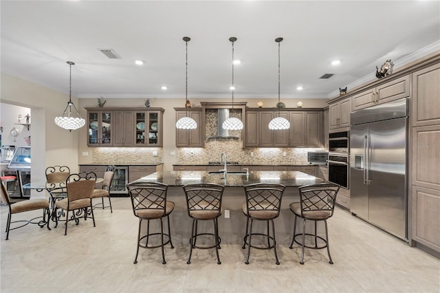 kitchen with sink, stainless steel built in fridge, an island with sink, pendant lighting, and wall chimney range hood