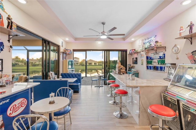 kitchen with a breakfast bar area, a water view, light wood-type flooring, a tray ceiling, and ceiling fan