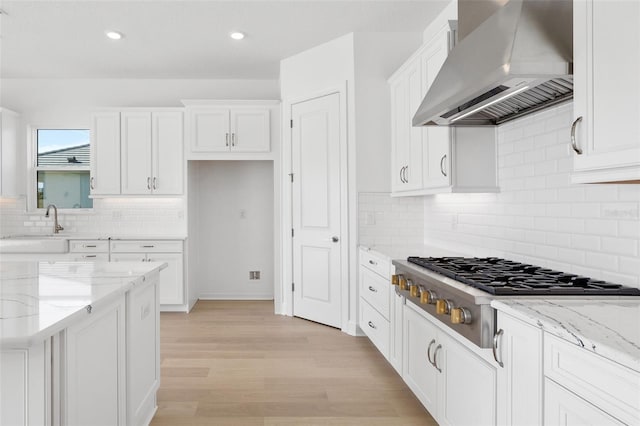 kitchen featuring wall chimney range hood, light stone countertops, light wood-type flooring, white cabinetry, and stainless steel gas cooktop