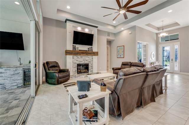 tiled living room featuring a tile fireplace, french doors, ceiling fan with notable chandelier, a raised ceiling, and beverage cooler