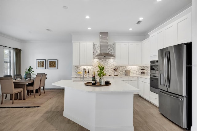 kitchen with an island with sink, stainless steel appliances, white cabinetry, and wall chimney range hood