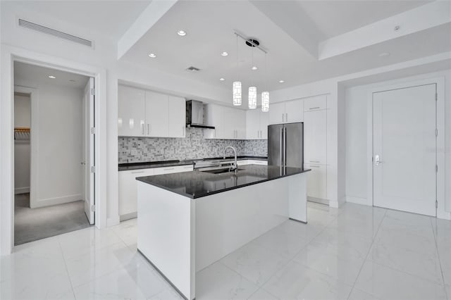 kitchen with high end fridge, an island with sink, pendant lighting, wall chimney range hood, and white cabinets