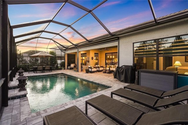 pool at dusk featuring a patio, outdoor lounge area, ceiling fan, and glass enclosure