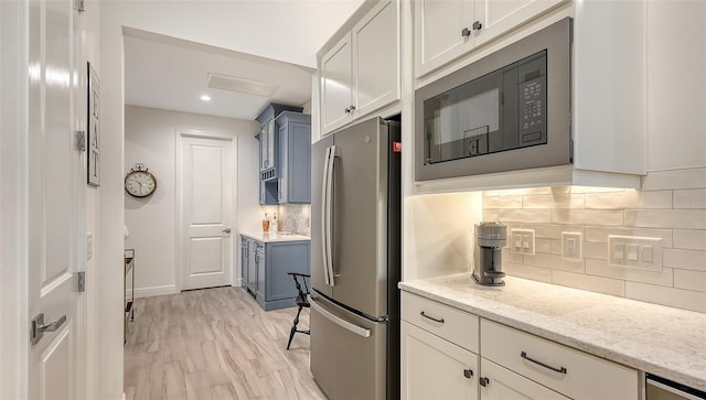 kitchen featuring light stone countertops, decorative backsplash, black microwave, light hardwood / wood-style floors, and stainless steel refrigerator