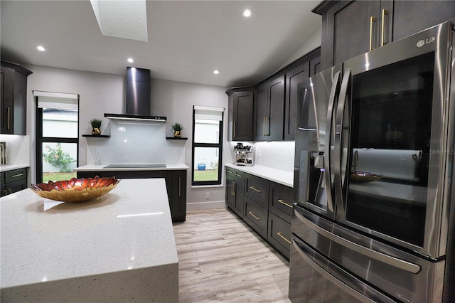 kitchen featuring light stone countertops, wall chimney exhaust hood, stainless steel refrigerator with ice dispenser, black electric stovetop, and light wood-type flooring