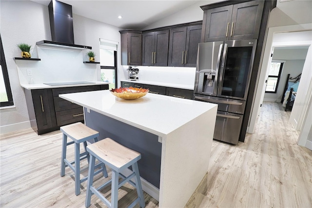 kitchen with stainless steel fridge, wall chimney exhaust hood, dark brown cabinetry, a kitchen island, and plenty of natural light