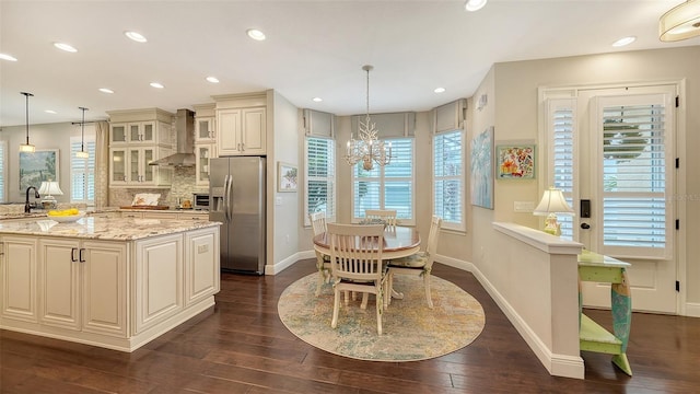 kitchen featuring stainless steel refrigerator with ice dispenser, wall chimney exhaust hood, light stone counters, and pendant lighting