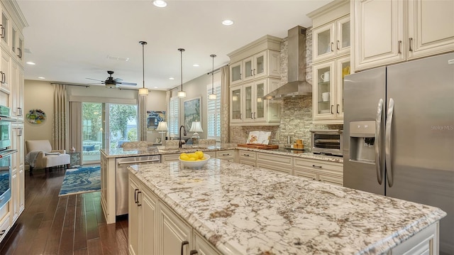 kitchen with wall chimney range hood, stainless steel appliances, sink, hanging light fixtures, and light stone counters