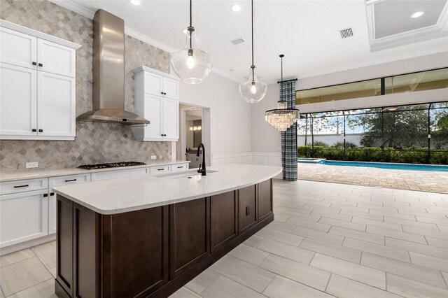 kitchen with white cabinetry, hanging light fixtures, a kitchen island with sink, and wall chimney range hood