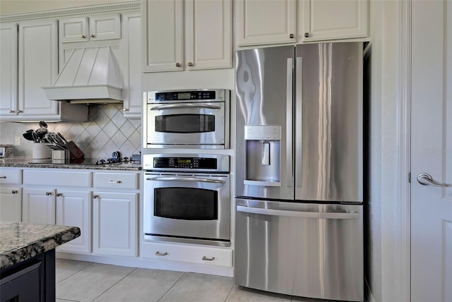 kitchen with appliances with stainless steel finishes, light tile patterned floors, dark stone counters, premium range hood, and white cabinets