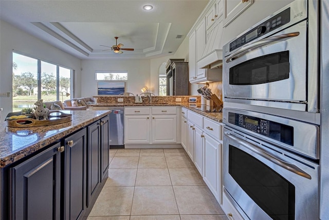 kitchen with kitchen peninsula, stainless steel appliances, a raised ceiling, white cabinetry, and dark stone countertops