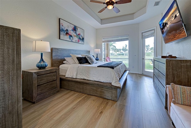 bedroom with ceiling fan, light hardwood / wood-style floors, a raised ceiling, and access to exterior