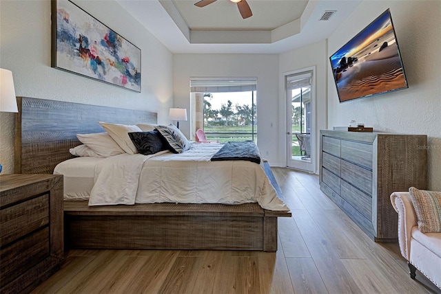 bedroom with ceiling fan, light hardwood / wood-style floors, access to outside, and a tray ceiling