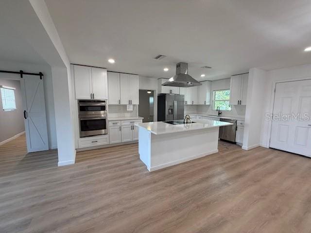 kitchen with white cabinetry, a barn door, appliances with stainless steel finishes, light wood-type flooring, and wall chimney exhaust hood