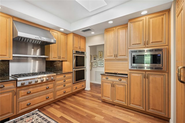 kitchen featuring a skylight, light hardwood / wood-style flooring, wall chimney exhaust hood, decorative backsplash, and stainless steel appliances