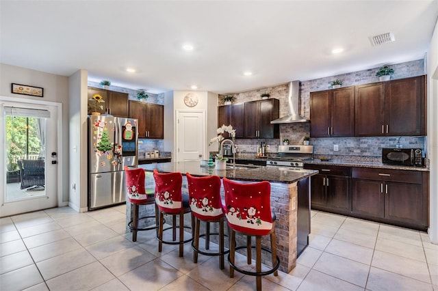 kitchen with stainless steel appliances, wall chimney range hood, dark stone countertops, a breakfast bar area, and an island with sink