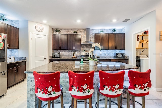 kitchen featuring dark brown cabinets, an island with sink, dark stone counters, and wall chimney range hood