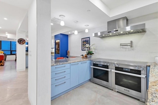 kitchen featuring wall chimney exhaust hood, blue cabinets, light stone counters, wall oven, and range with two ovens