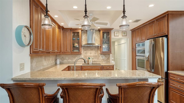 kitchen featuring a raised ceiling, hanging light fixtures, wall chimney exhaust hood, a kitchen bar, and stainless steel appliances